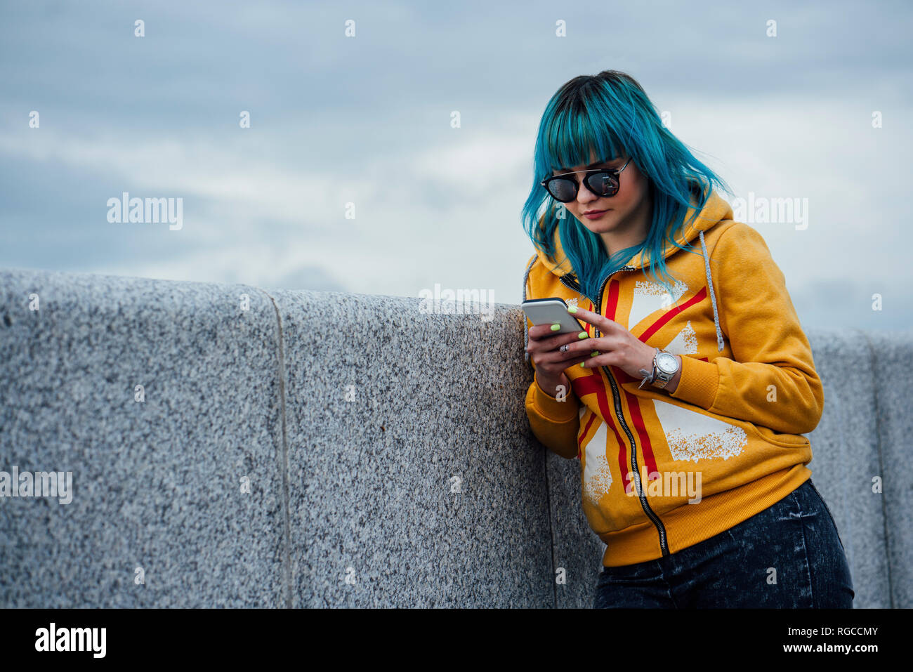 Portrait of young woman with dyed blue hair leaning against wall looking at cell phone Stock Photo