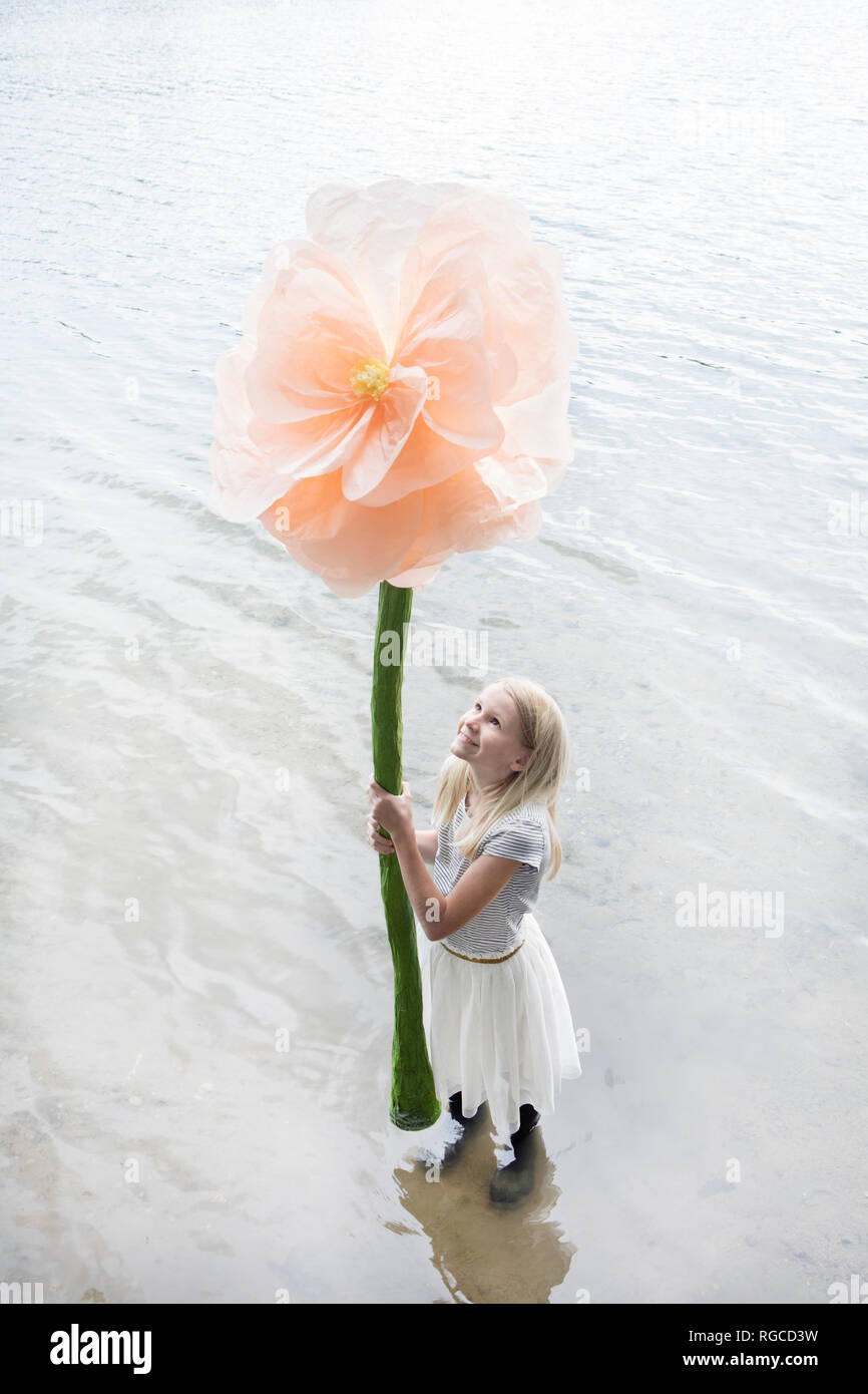 Smiling blond girl standing in a lake holding oversized artificial flower Stock Photo