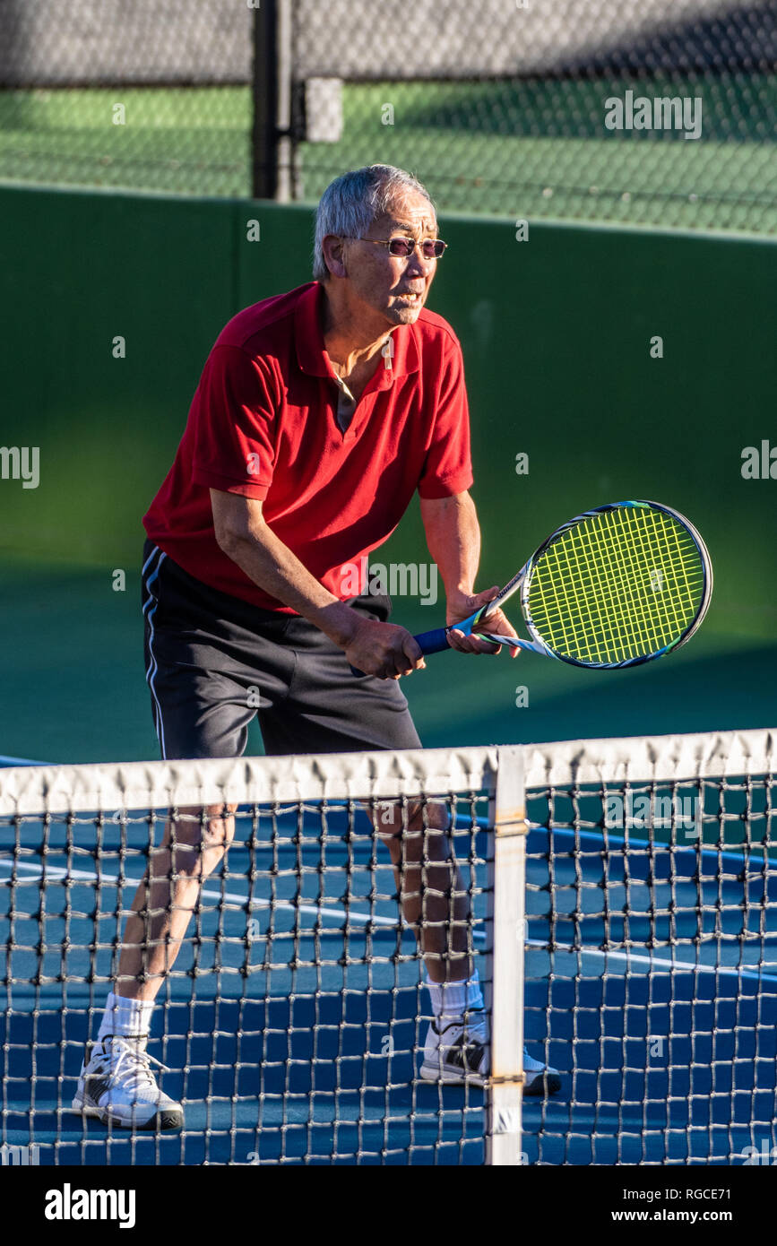 Focused Chinese elderly man ready with racquet raised during a game of tennis. Stock Photo