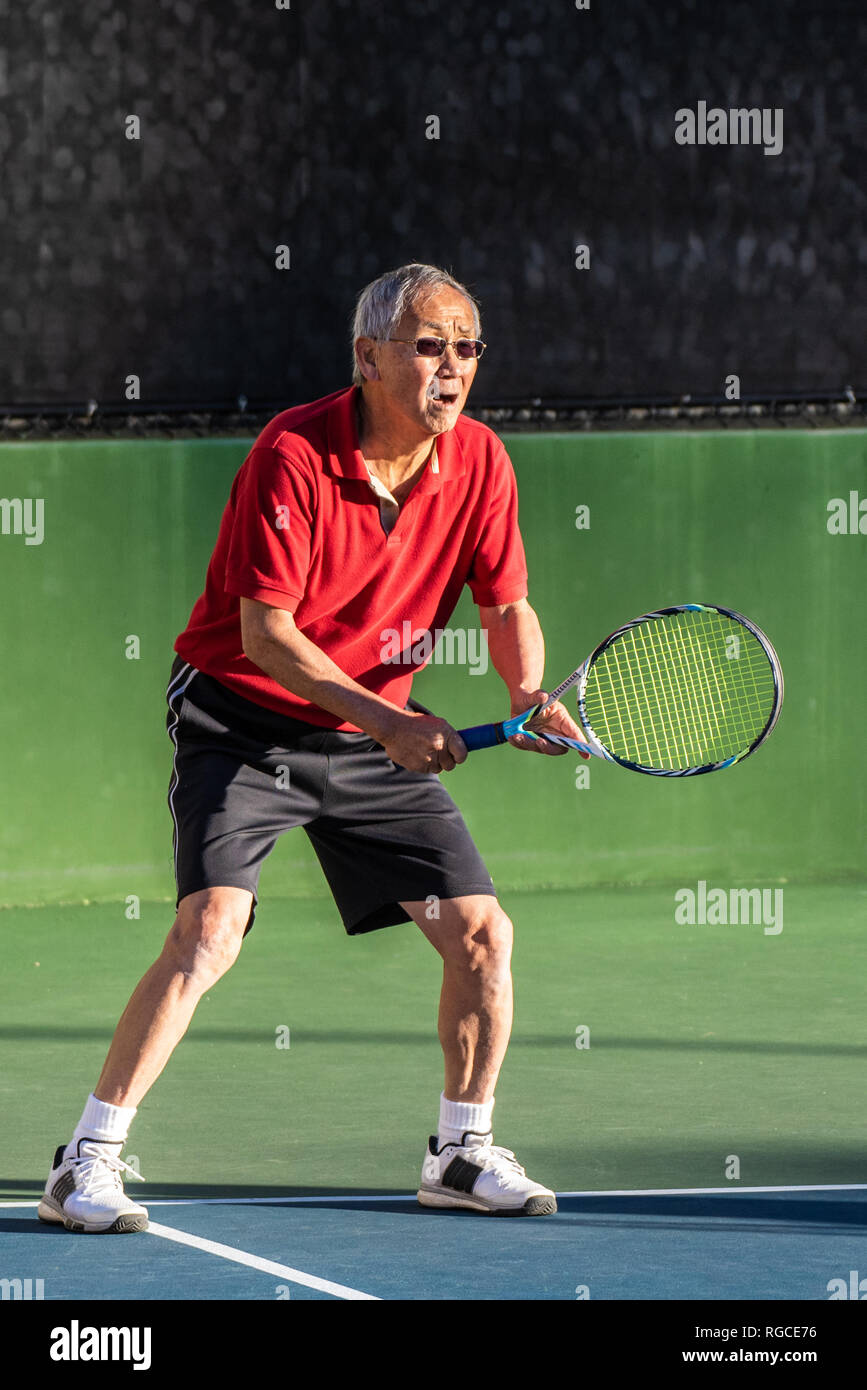 Focused Chinese elderly man ready with racquet raised and serious expression during a game of tennis. Stock Photo