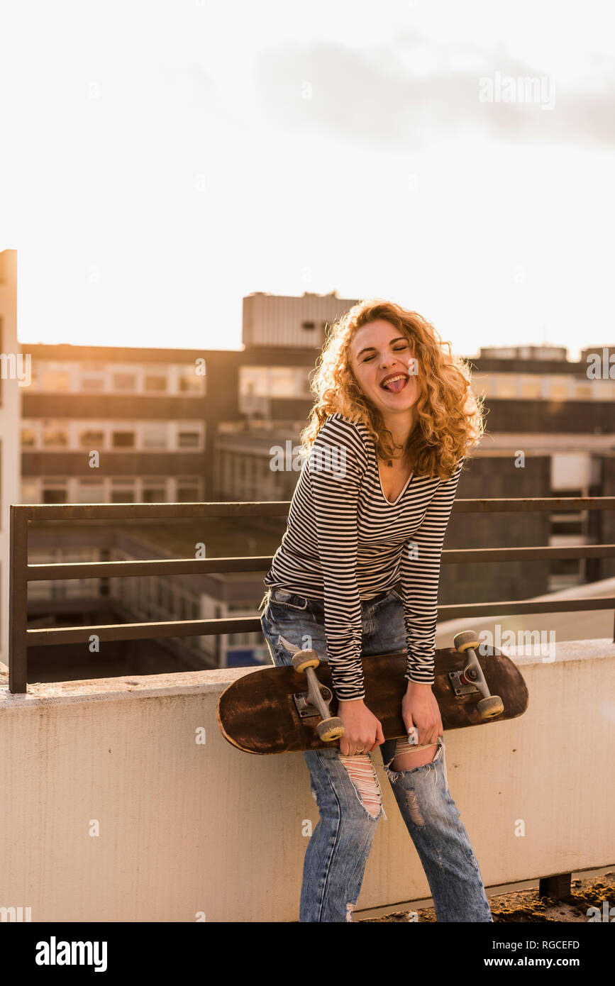 Portrait of cheeky young woman with skateboard on roof terrace at sunset Stock Photo