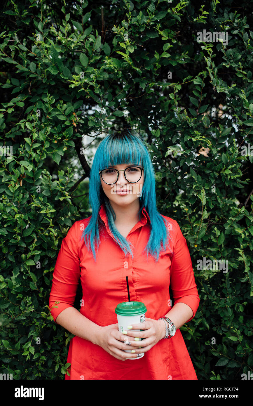 Portrait of young woman with dyed blue hair standing in front of a hedge with beverage Stock Photo