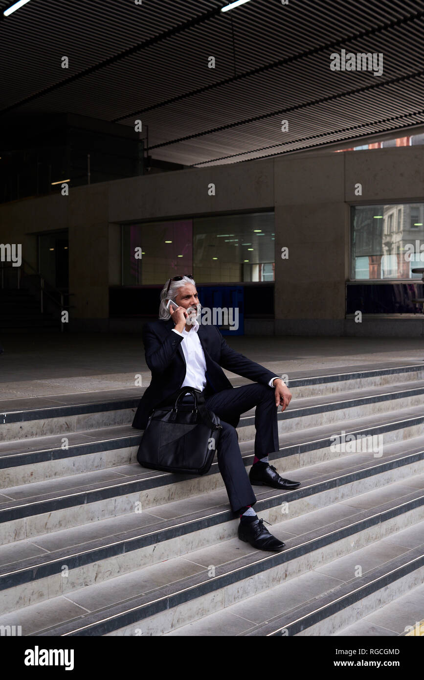 Stylish senior businessman on the phone sitting on stairs outdoors Stock Photo