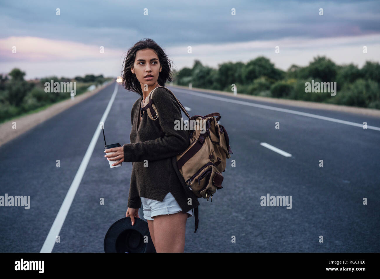 Portrait of young hitchhiking woman with backpack and beverage standing on lane Stock Photo