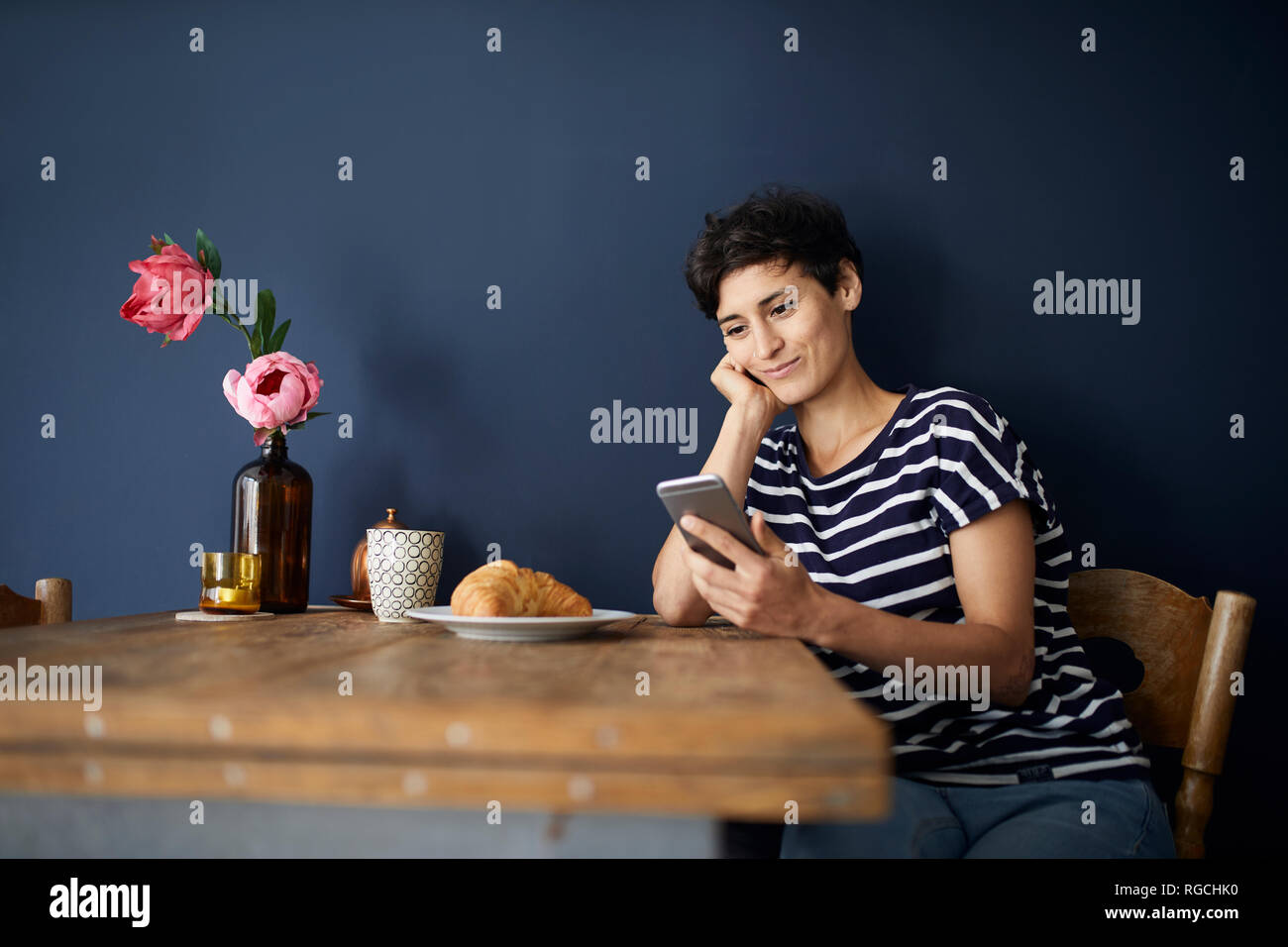 Smiling woman at home sitting at wooden table checking cell phone Stock Photo