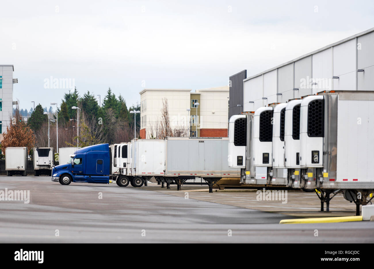 Big rig blue semi truck and refrigerated semi trailers standing in row in warehouse dock for loading and unloading commercial cargo and continuing go  Stock Photo
