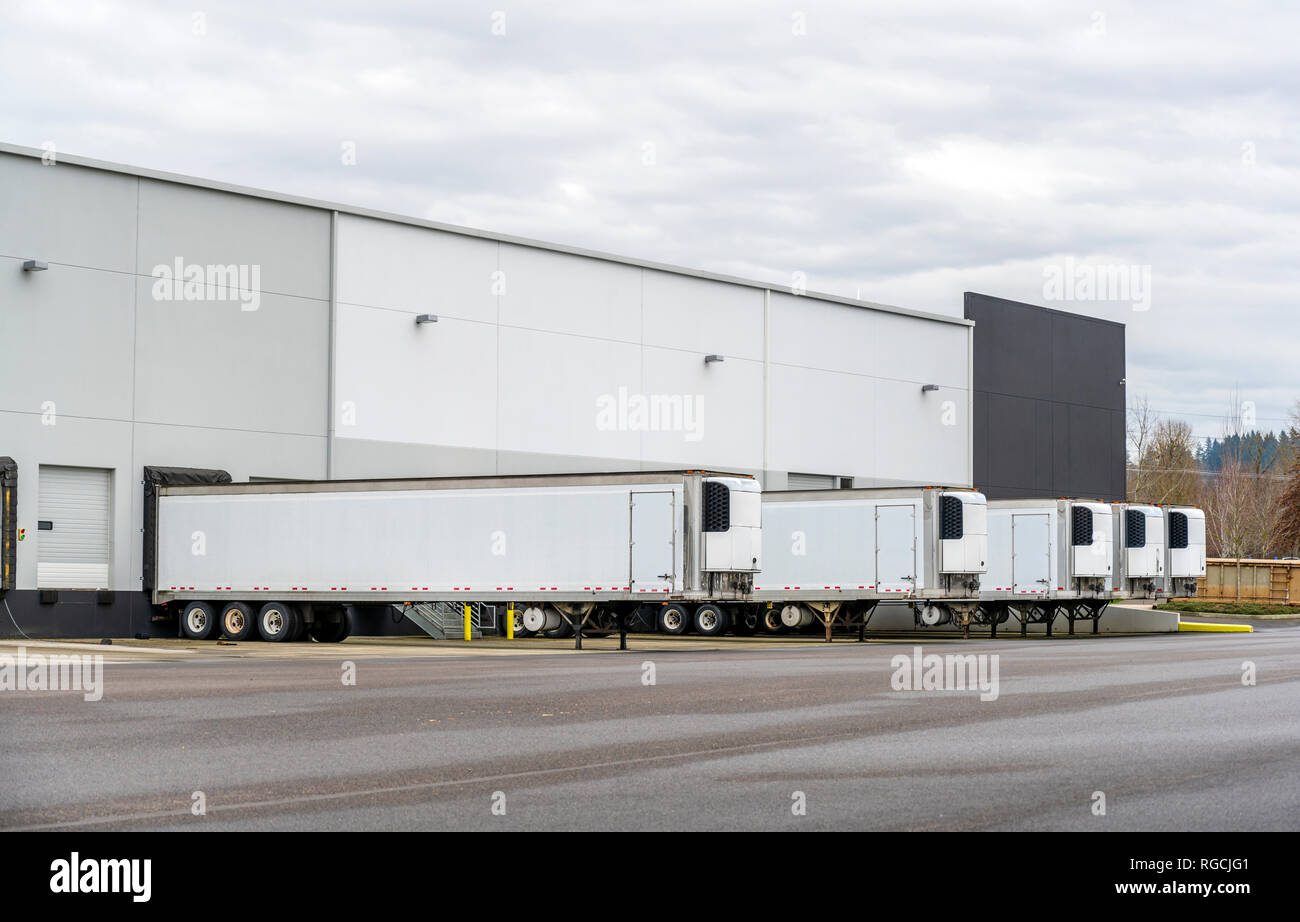 Long refrigerator semi trailers stand in row in warehouse docks with gates and loading and unloading commercial cargo and continuing go to the destina Stock Photo