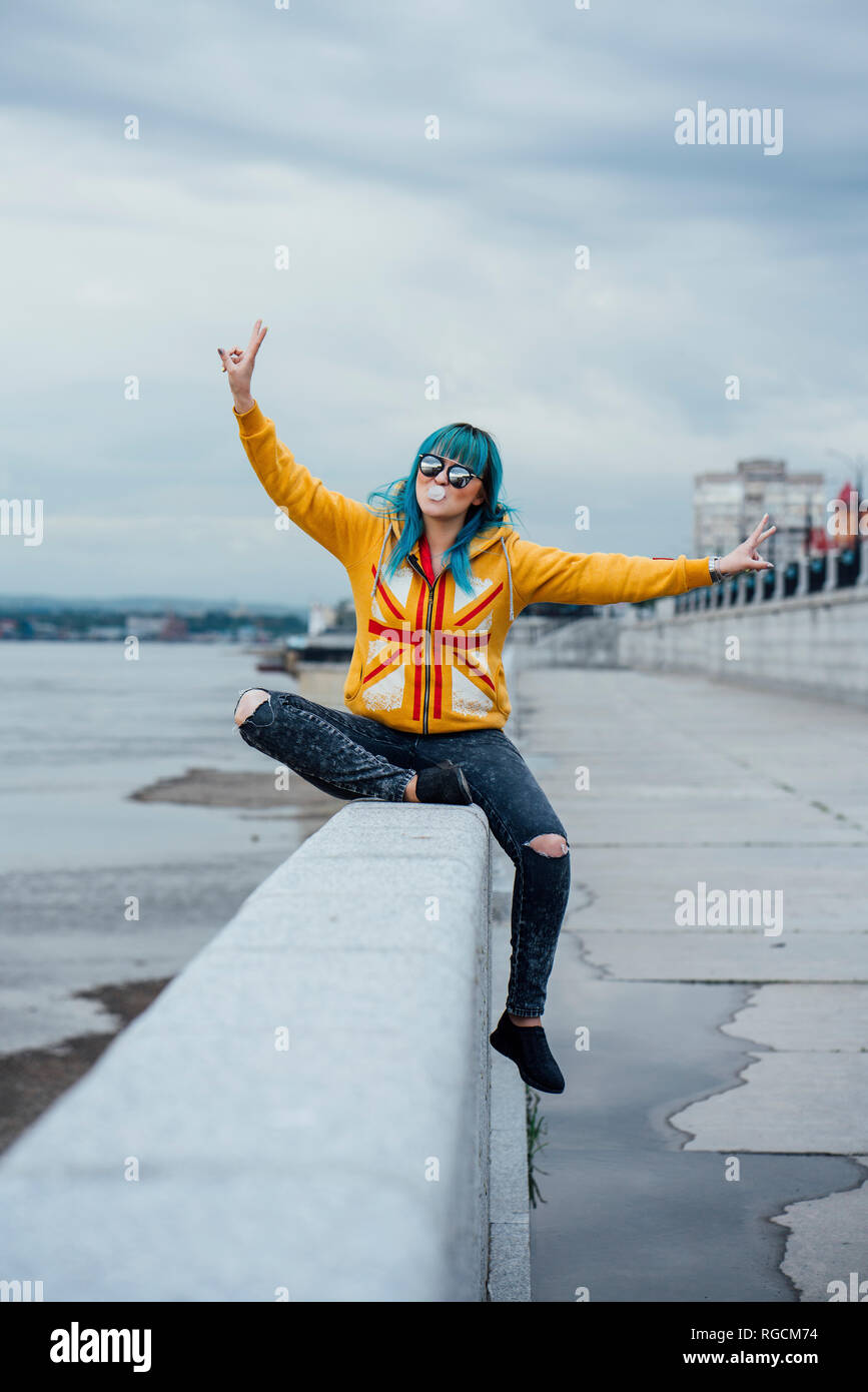 Portrait of young woman with dyed blue hair sitting on a wall showing victory signs Stock Photo