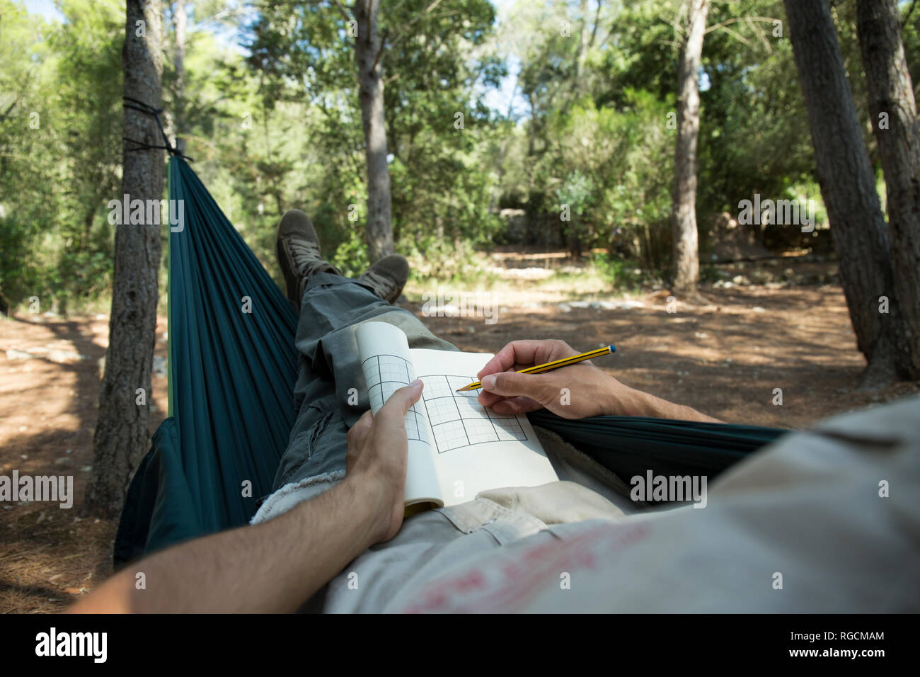 Man lying in hammock in the woods relaxing with puzzle book, partial view Stock Photo