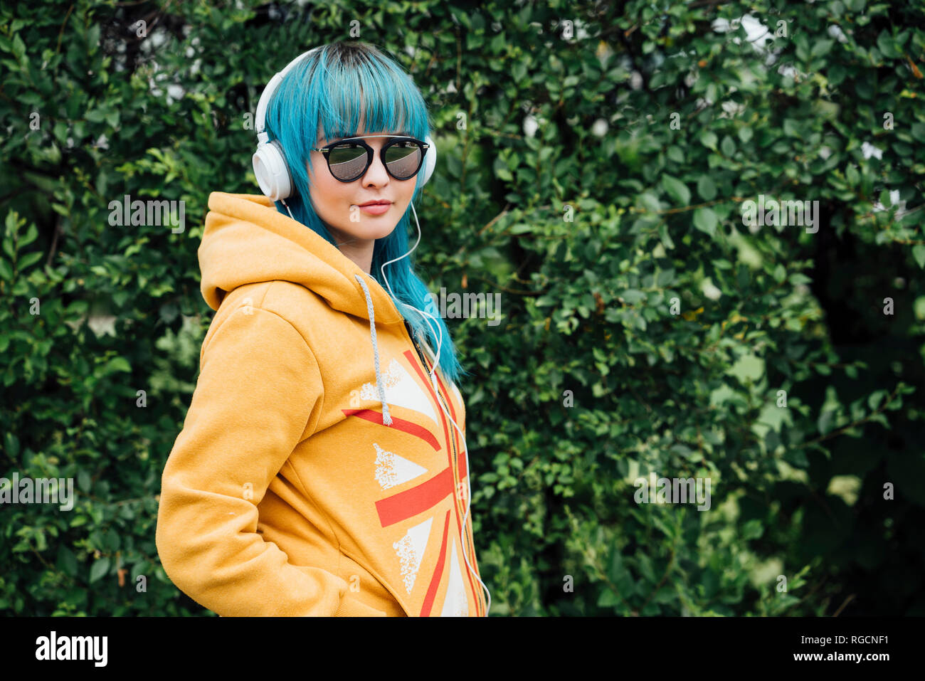 Portrait of young woman with dyed blue hair and headphones wearing yellow hooded jacket Stock Photo