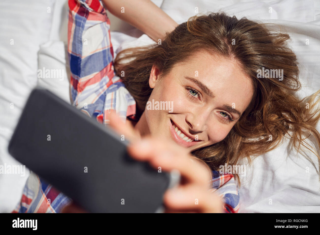 Portrait of laughing woman lying on bed taking selfie with smartphone Stock Photo