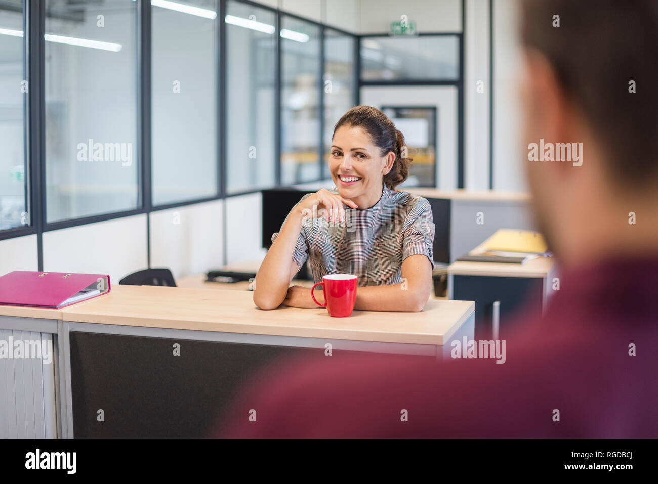 Female employee sitting at desk, talking to colleague Stock Photo