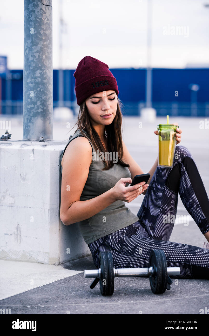 Young woman sitting on ground after dumbbell training, using smartphone Stock Photo