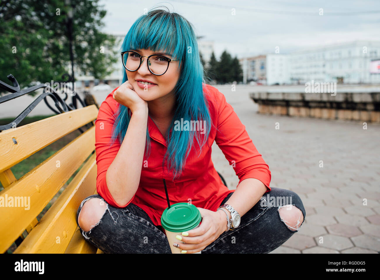 Portrait of smiling young woman with dyed blue hair sitting on a bench with beverage Stock Photo