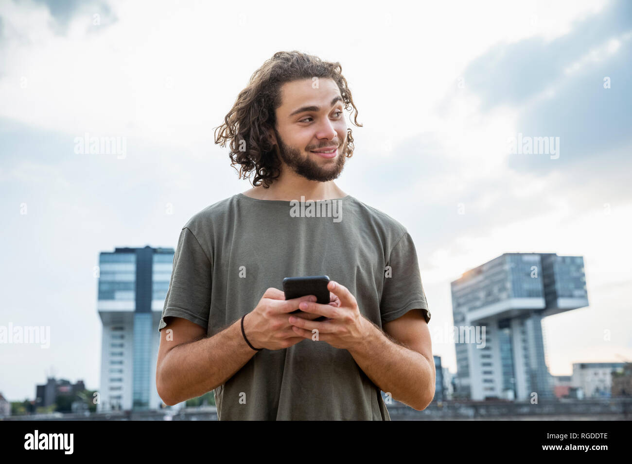 Germany, Cologne, smiling young man holding cell phone looking sideways Stock Photo