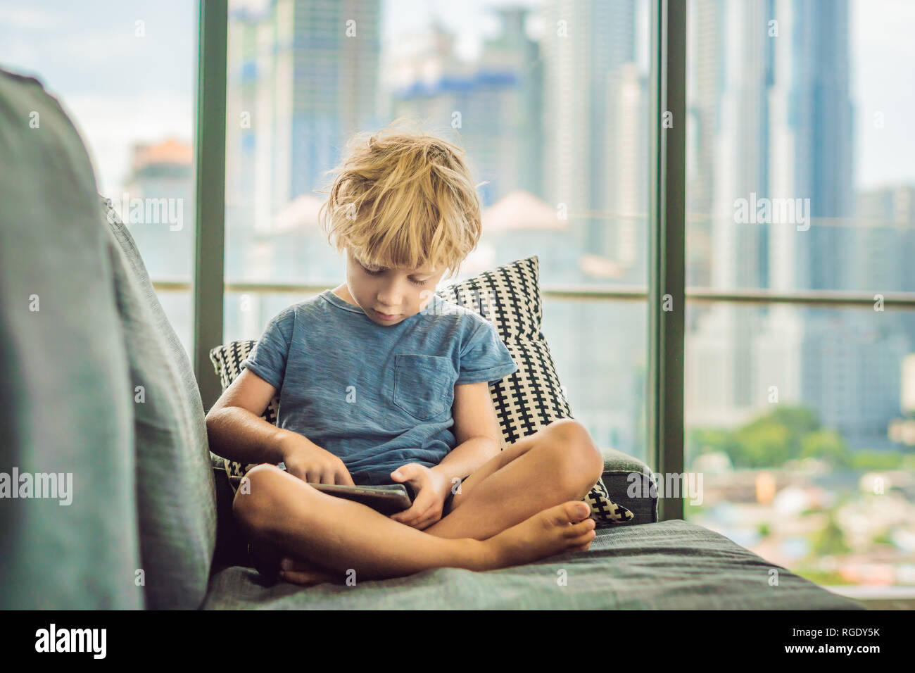 Boy uses a tablet at home on the couch in the background of a window with skyscrapers. Modern children in the megalopolis use a tablet concept Stock Photo