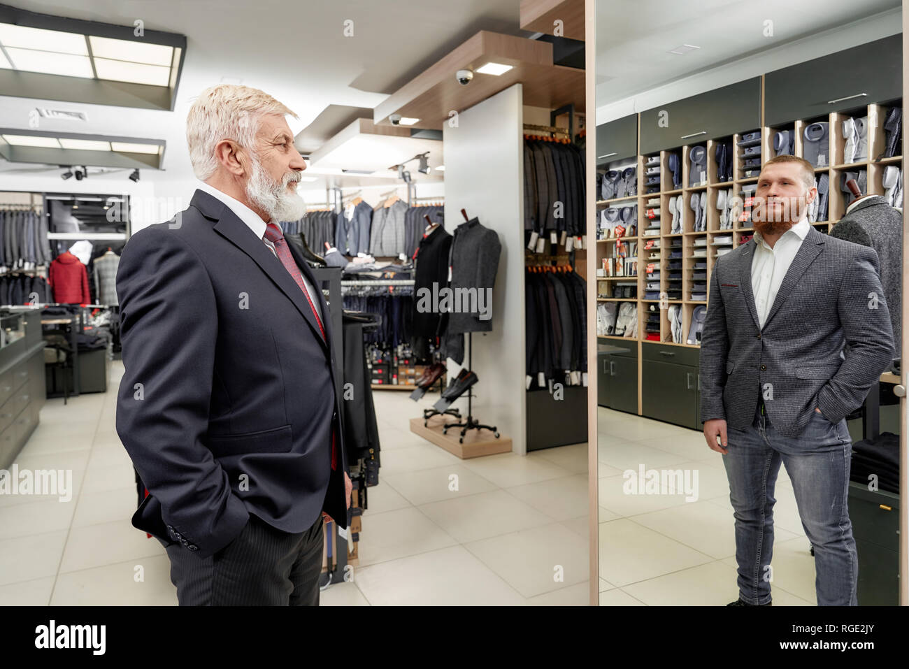 Elderly, bearded man with grey hair trying on classic suit with tie and shirt in boutique. Man looking at mirror and seeing reflection of young trendy and charming man. Expensive menswear garment. Stock Photo