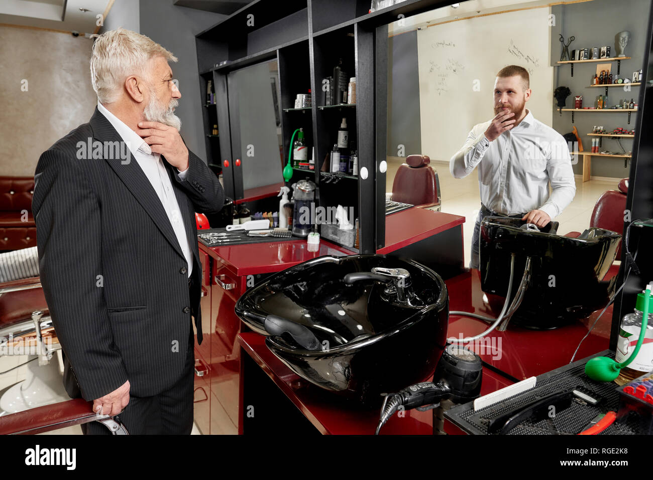 Pensioner standing in barbershop near dressing table. Man holding hand on chin and observing reflection of young charming man. Elderly man wearing in black classic suit and having grey hair. Stock Photo