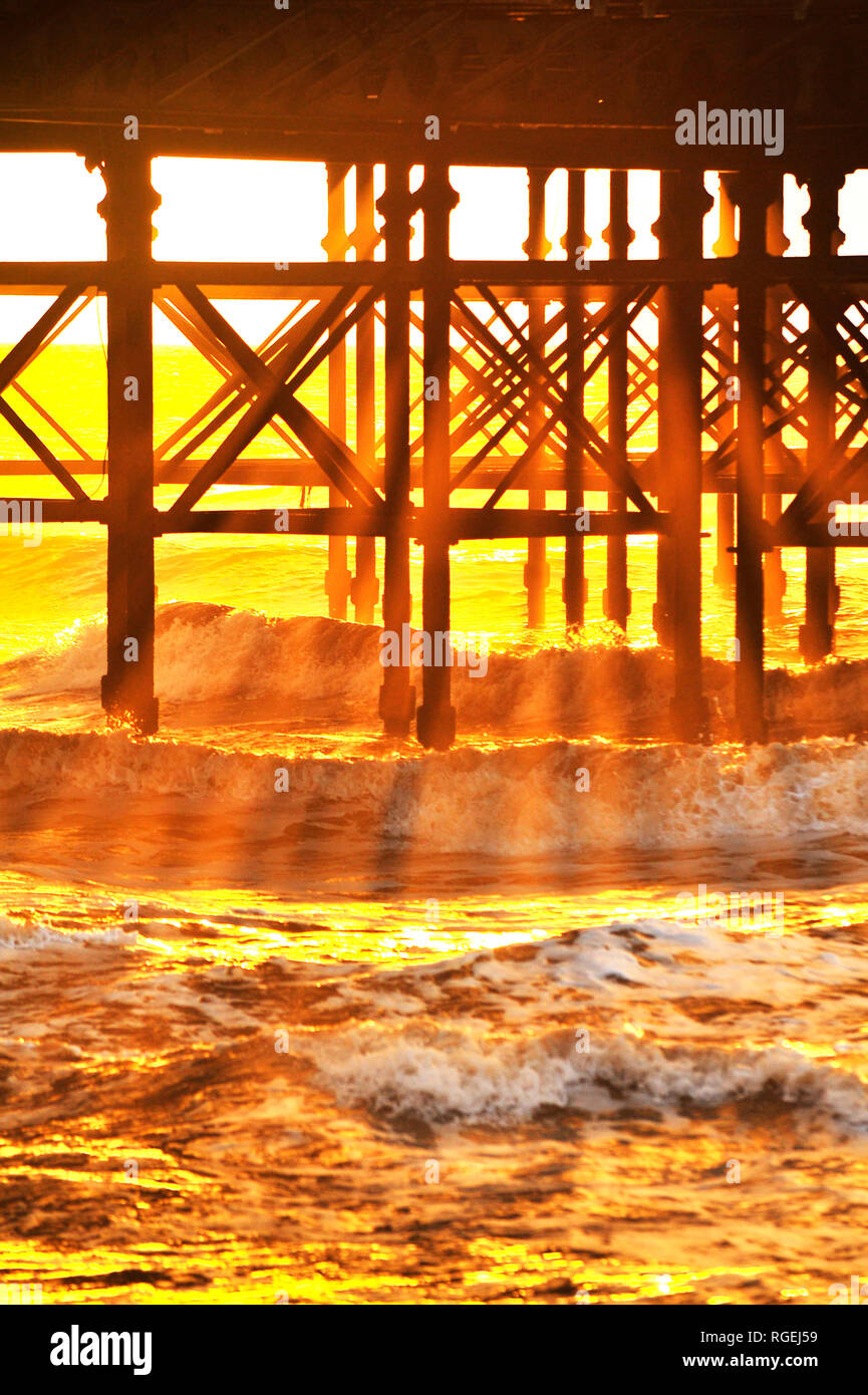 Golden shafts of light pass through the iron work underneath South Pier and into the waves of the incoming tide Stock Photo