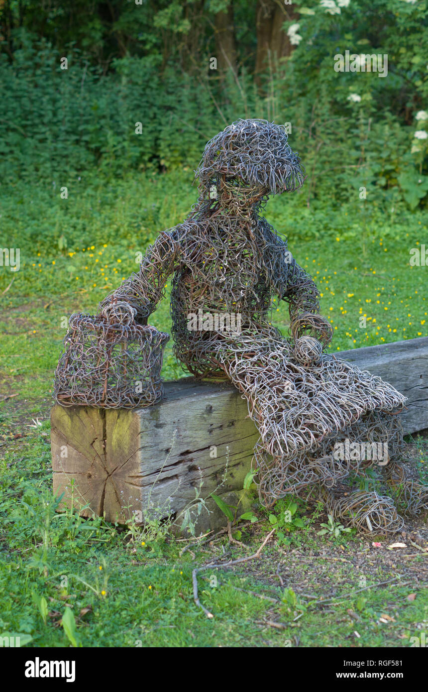 Sculpture of a woman with a shopping bag, made of metal wire, sitting on a wooden sleeper by the canal, Rothersthorpe, Northamptonshire, UK Stock Photo
