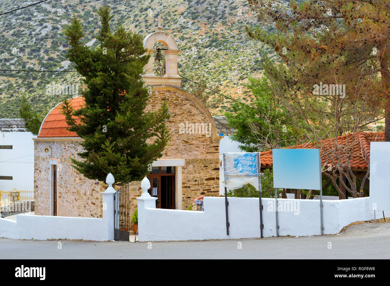 Eastern Orthodox Church, religious building made of stone with red tiled roof and bell in arch. Ekklisia Agia Triada. On background is mountains. Resort village Bali, Crete island Greece Stock Photo