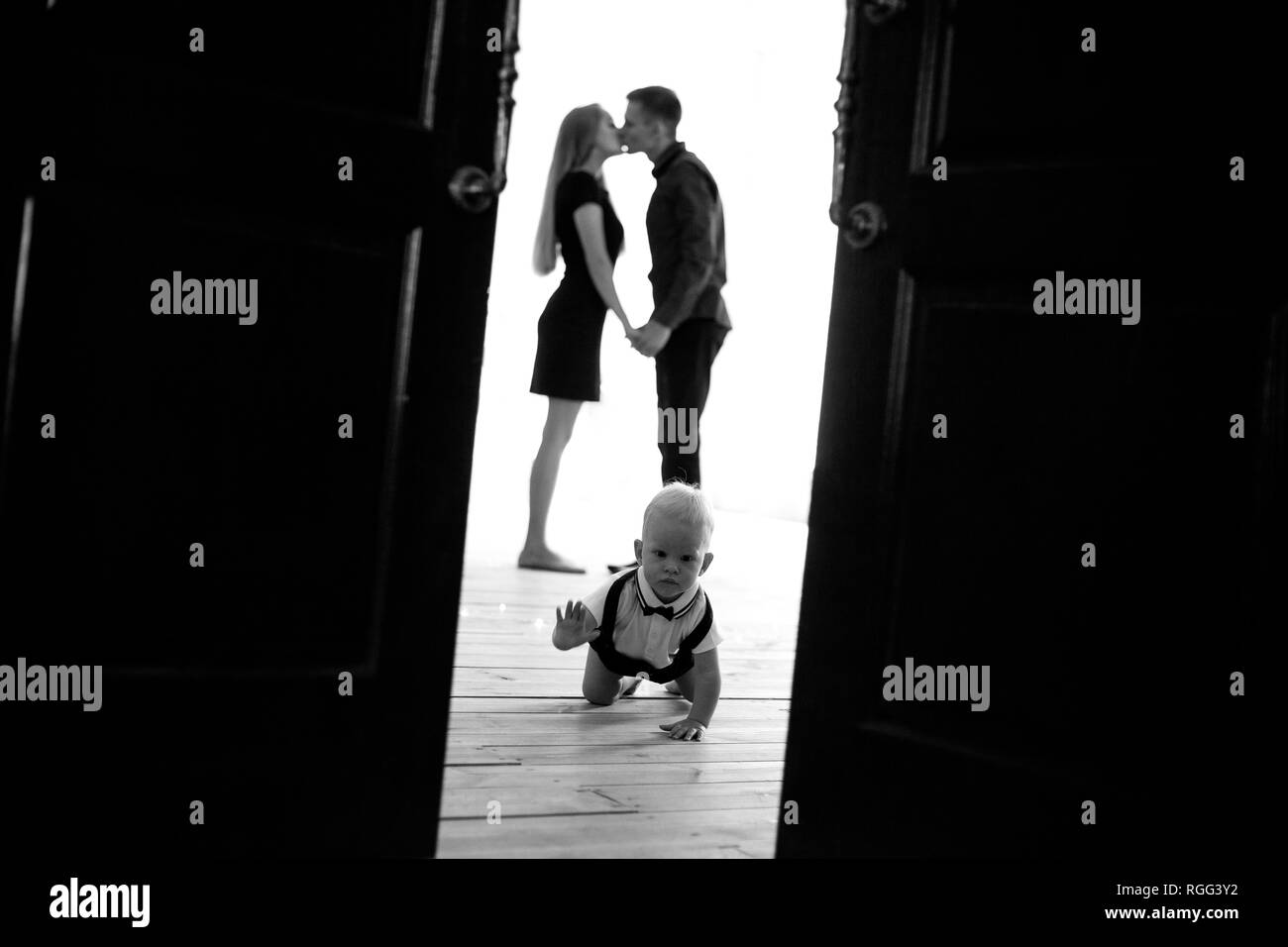 Baby boy crawls at the floor on background of his kissing parents. View through the opened door. Black and white image. Stock Photo