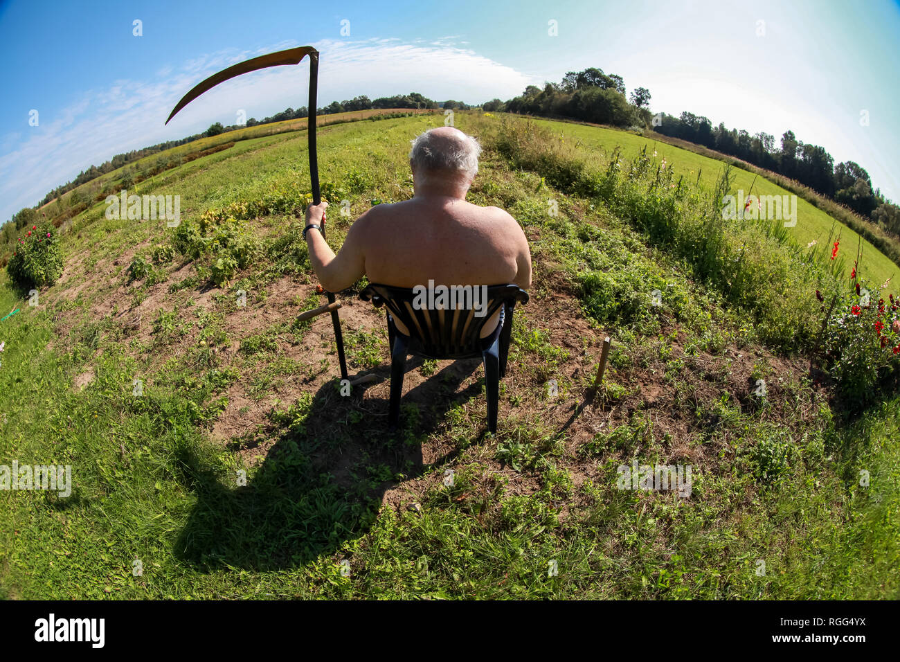 Farmer with a scythe sitting in the meadow. Landscape with old farmer sitting in green meadow. Farmer, meadow and blue sky. Stock Photo