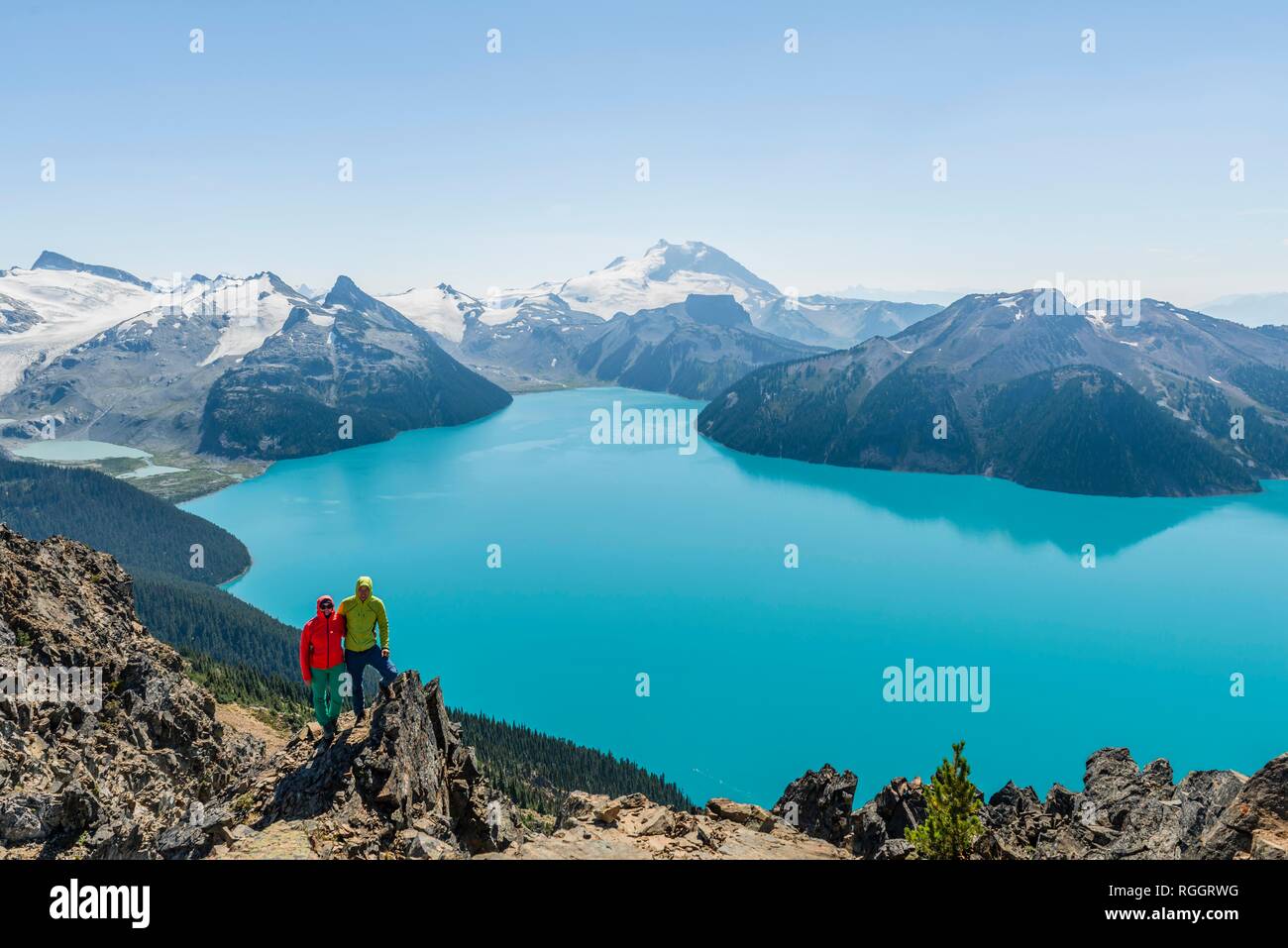 View from Panorama Ridge trail, two hikers on a rock, Garibaldi Lake, turquoise glacial lake, Guard Mountain and Deception Peak Stock Photo