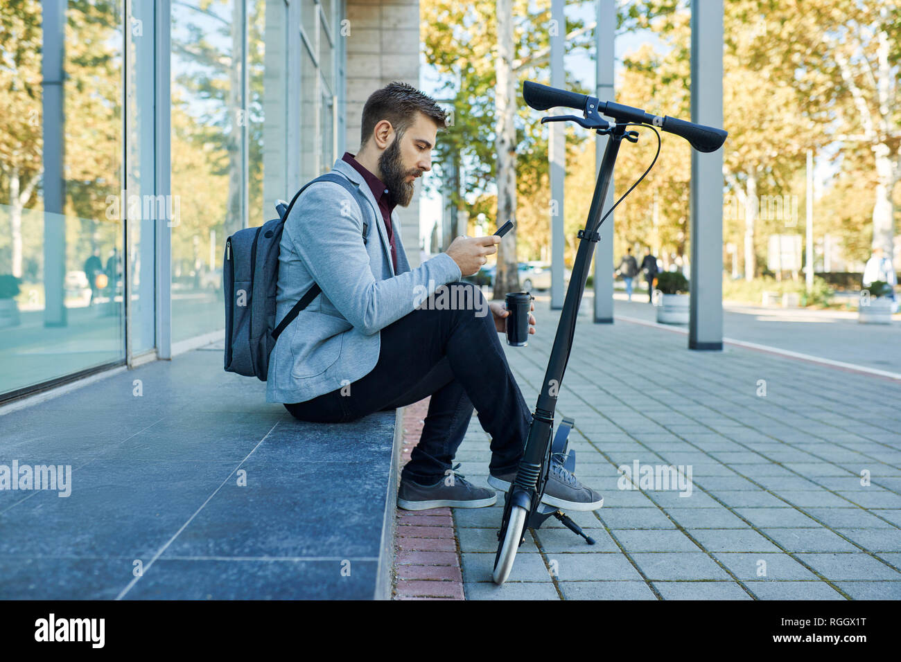 Businessman sitting outdoors using cell phone next to scooter Stock Photo