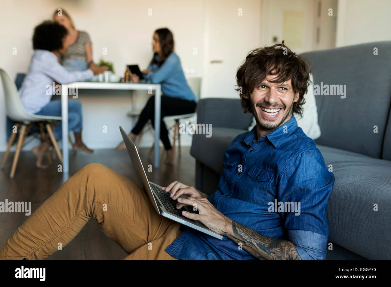 Laughing man sitting on floor using laptop with friends in background Stock Photo
