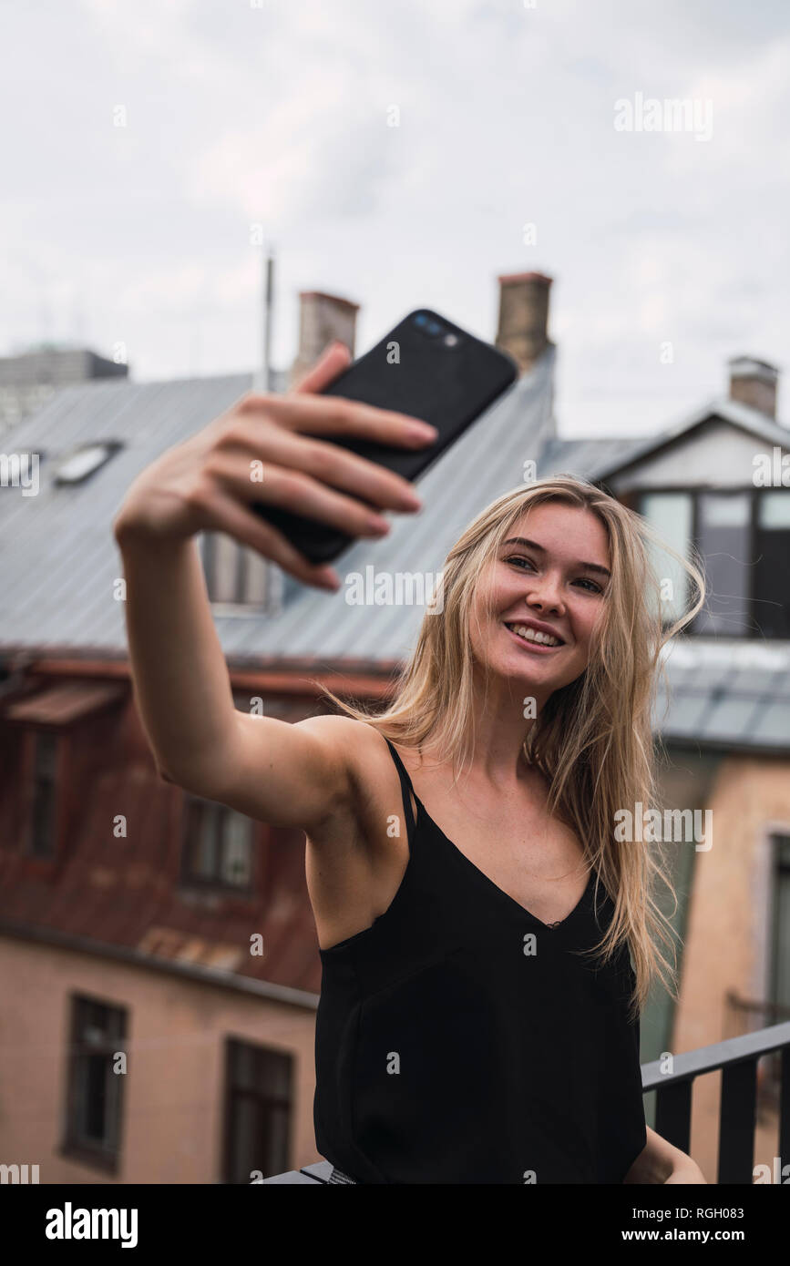 Portrait of smiling blond woman taking selfie on roof terrace Stock Photo