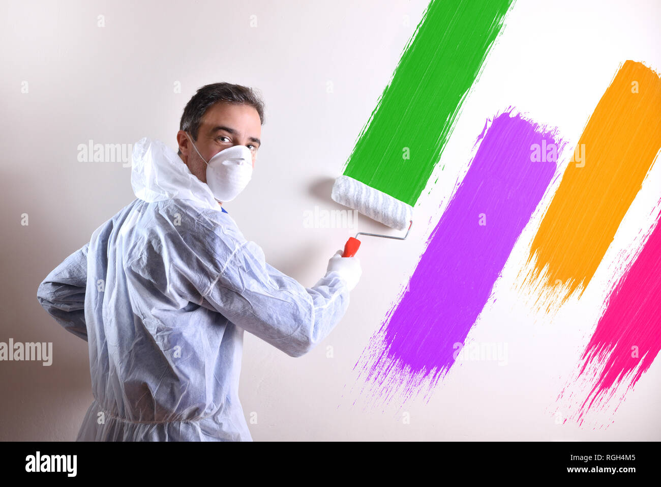 Background with professional painter with working overalls and roller on the back of a white wall painted with four colors. Horizontal composition. Stock Photo