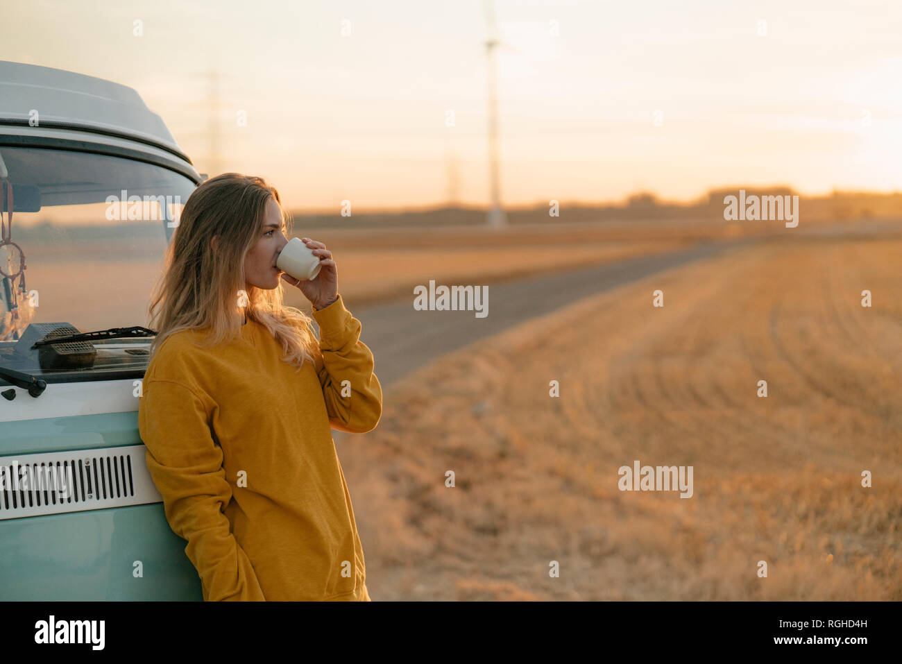 Young woman drinking from mug at camper van in rural landscape at sunset Stock Photo