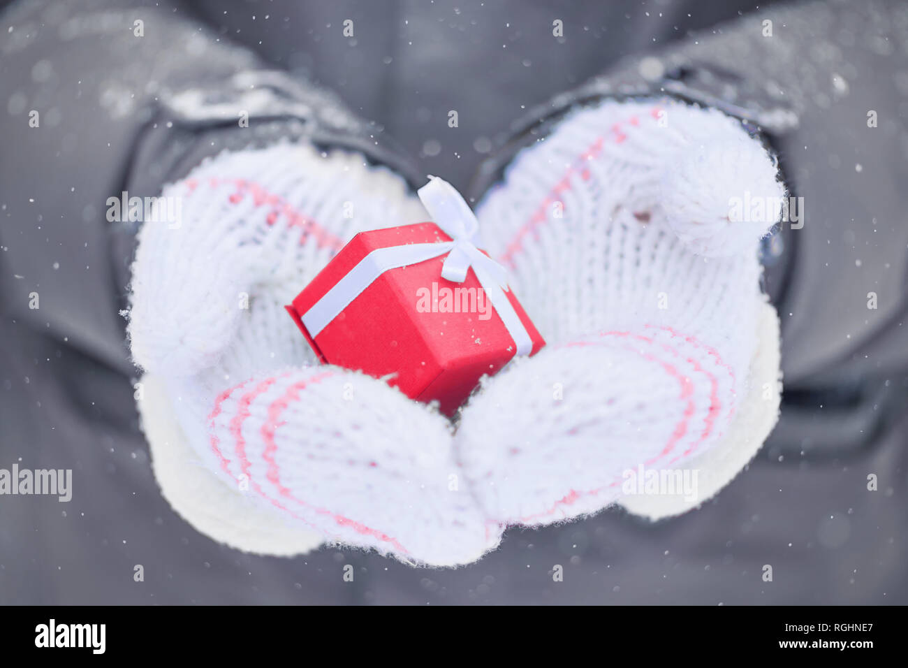 two hands in white mittens holding a small red box with a present outside on snowy winter day Stock Photo