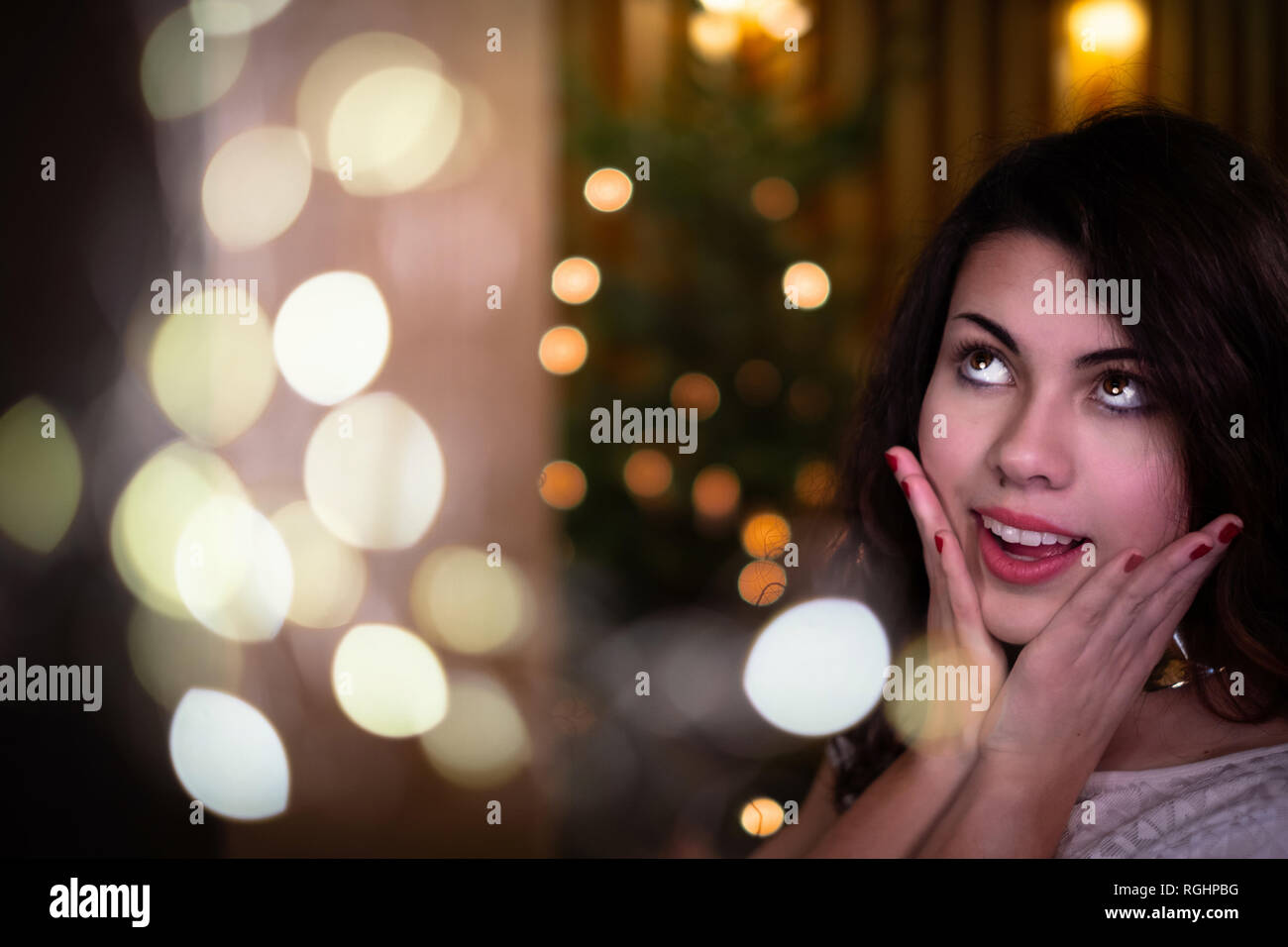 Beautiful young woman in front of christmas tree looking up in belief. Stock Photo