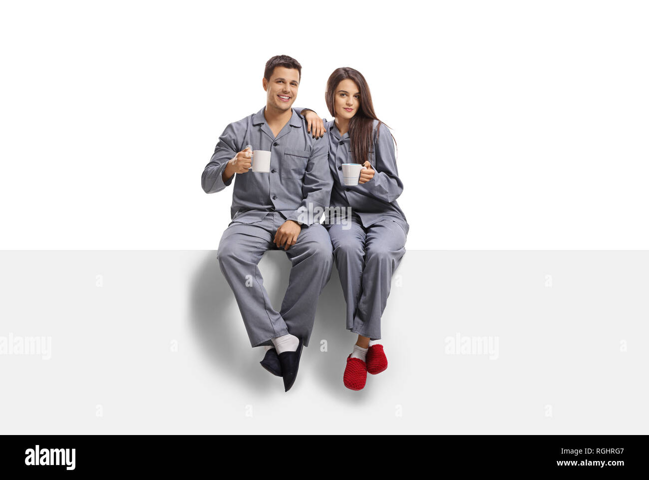Full length portrait of a young male and female in pyjamas and sleepers sitting on a white panel and holding coffee mugs isolated on white background Stock Photo