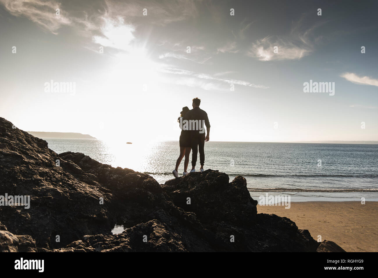 France, Brittany, rear view of young couple standing on rock at the beach at sunset Stock Photo