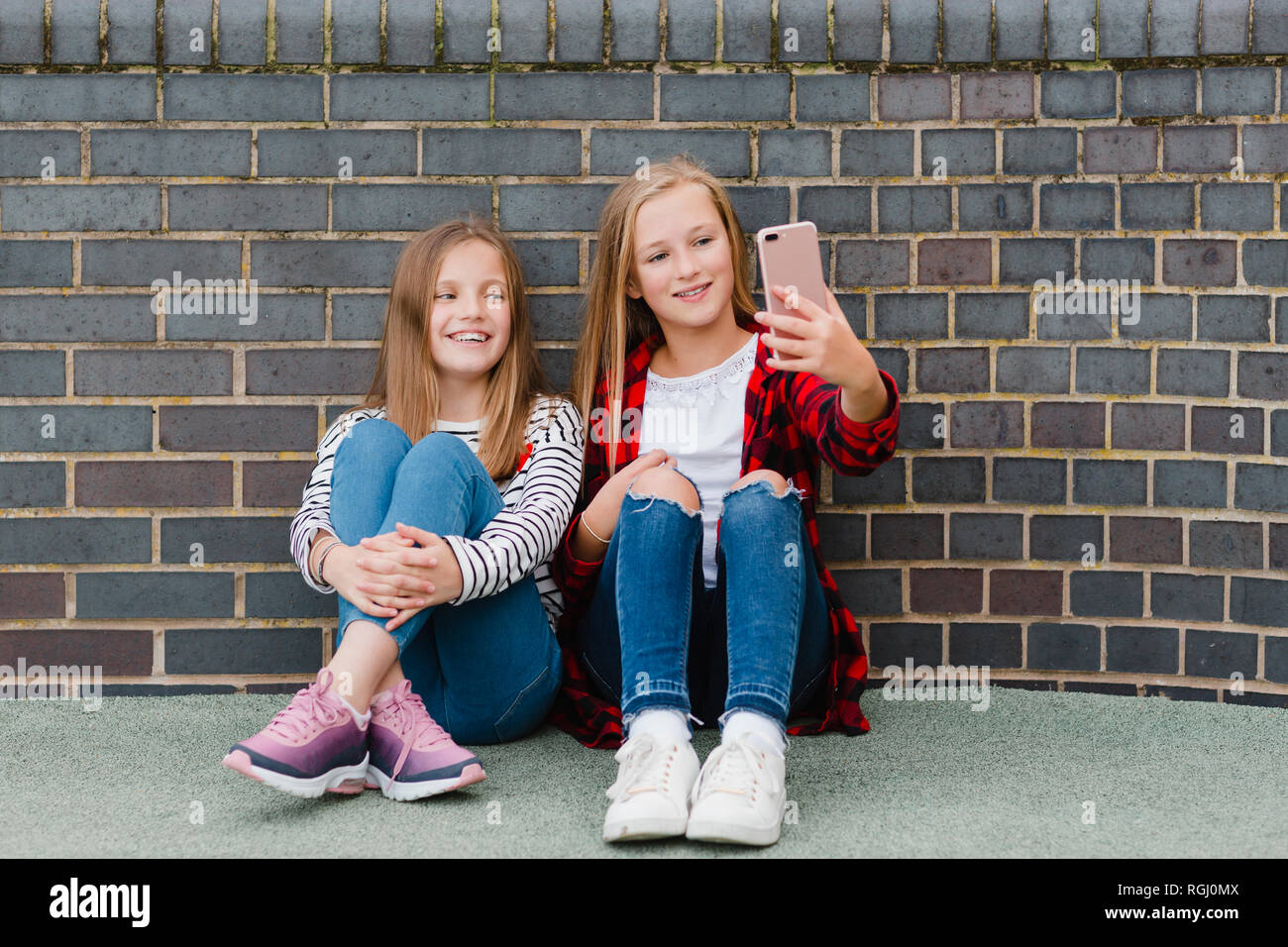 Portrait of two smiling girls sitting in front of brick wall taking selfie with smartphone Stock Photo