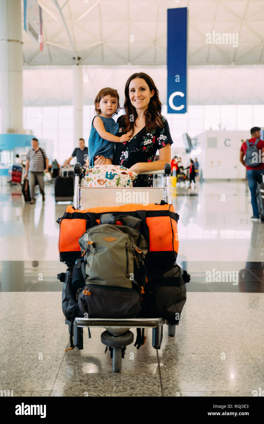Mother holding a baby girl at the airport and pushing a trolley with the luggage Stock Photo