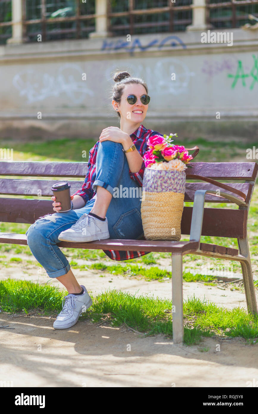 Front view of a young hipster Woman sitting on a park bench relaxing in ...