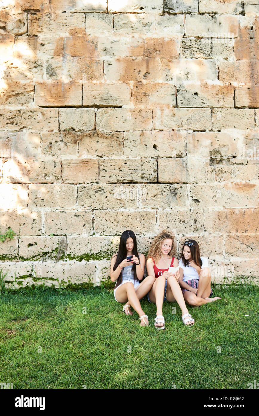 Three young women sitting at stone wall using cell phones listening to music Stock Photo