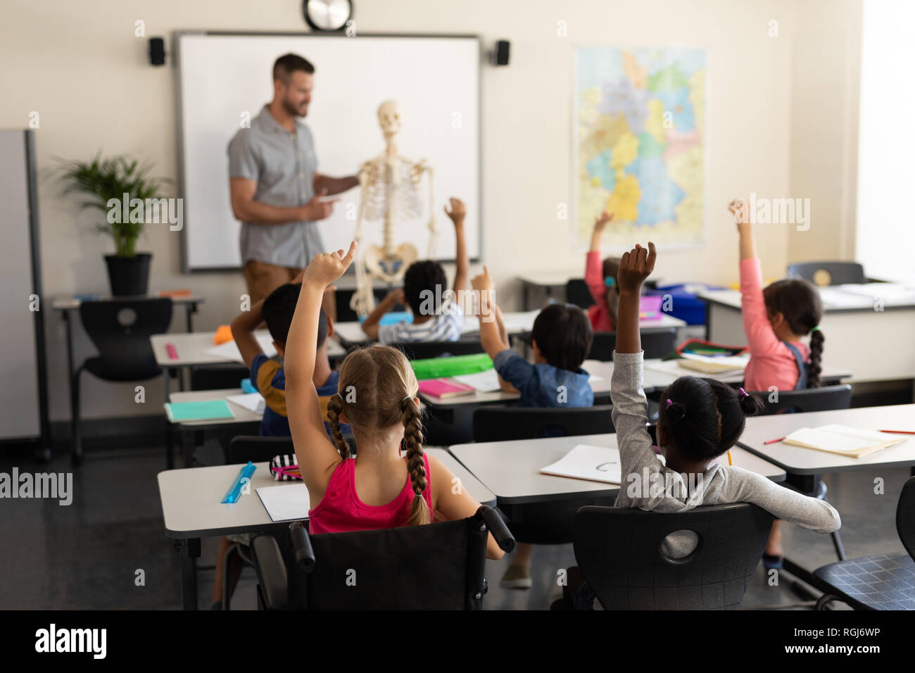 Rear view of school kids raising hand and sitting at desk in elementary ...