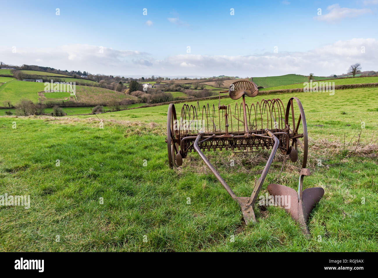 Old rusty horse drawn hay rake in a grass field with Devonshire countryside and hills in the background under a blue and cloudy sky on a bright day Stock Photo