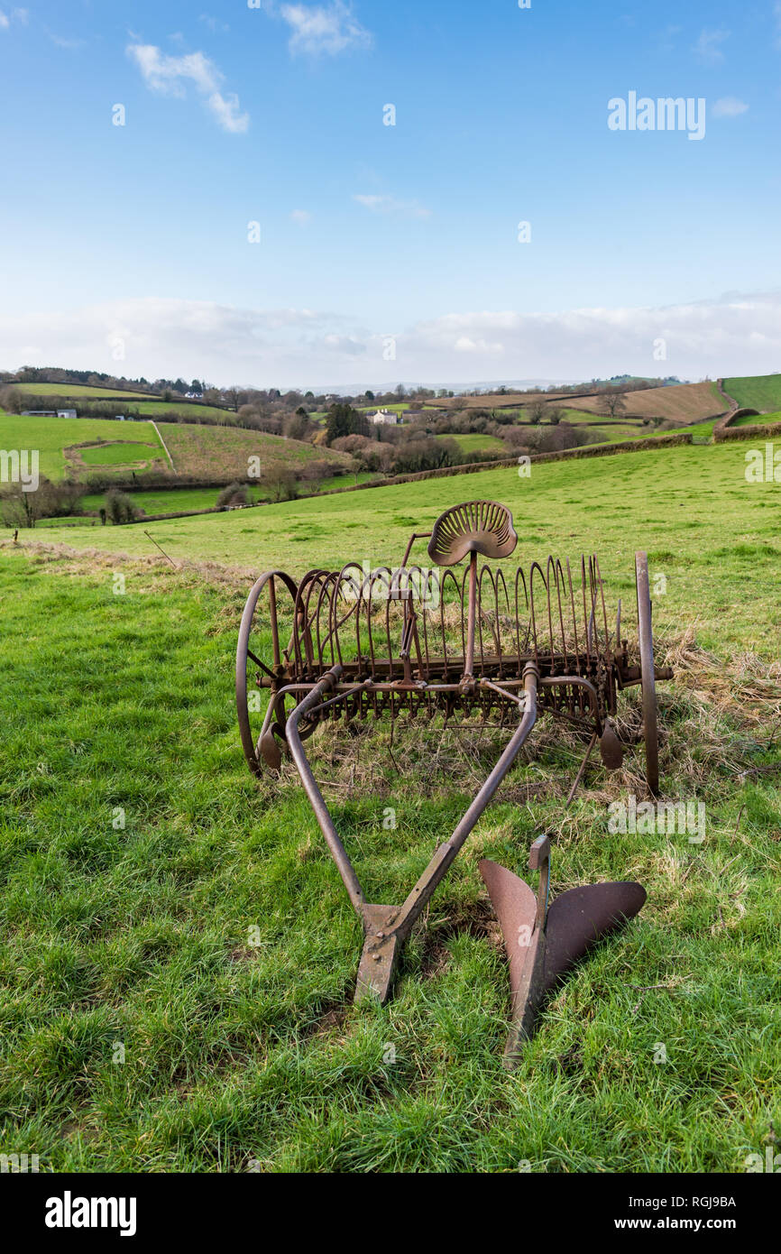 Old rusty horse drawn hay rake in a grass field with Devonshire countryside and hills in the background under a blue and cloudy sky on a bright day Stock Photo