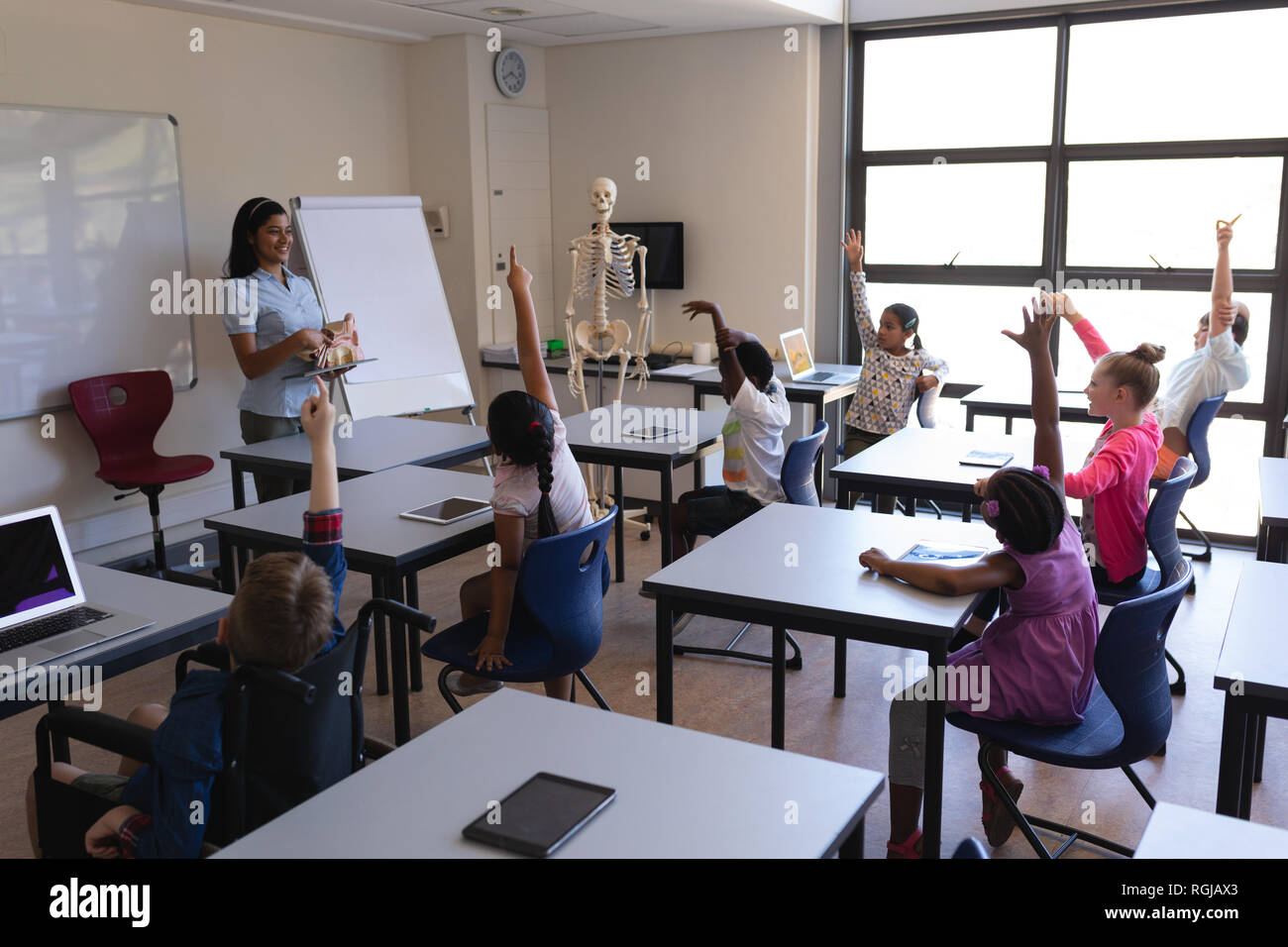 Side view of schoolkids raising hands while sitting at desk in ...