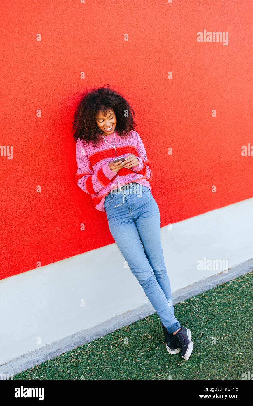 Smiling young woman with earphones leaning against red wall looking at cell phone Stock Photo
