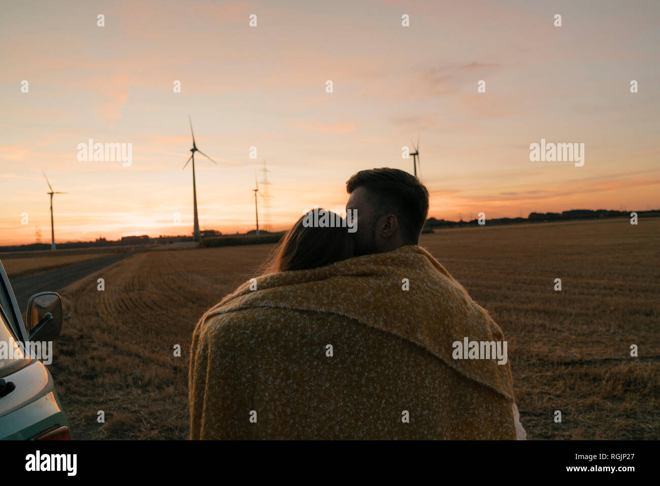 Couple wrapped in a blanket at camper van in rural landscape with wind turbines in background