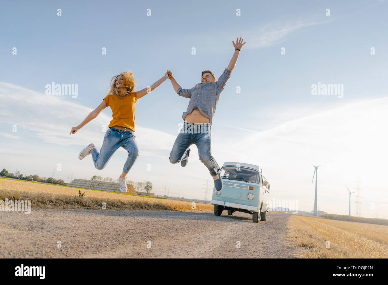 Exuberant couple jumping on dirt track at camper van in rural landscape Stock Photo