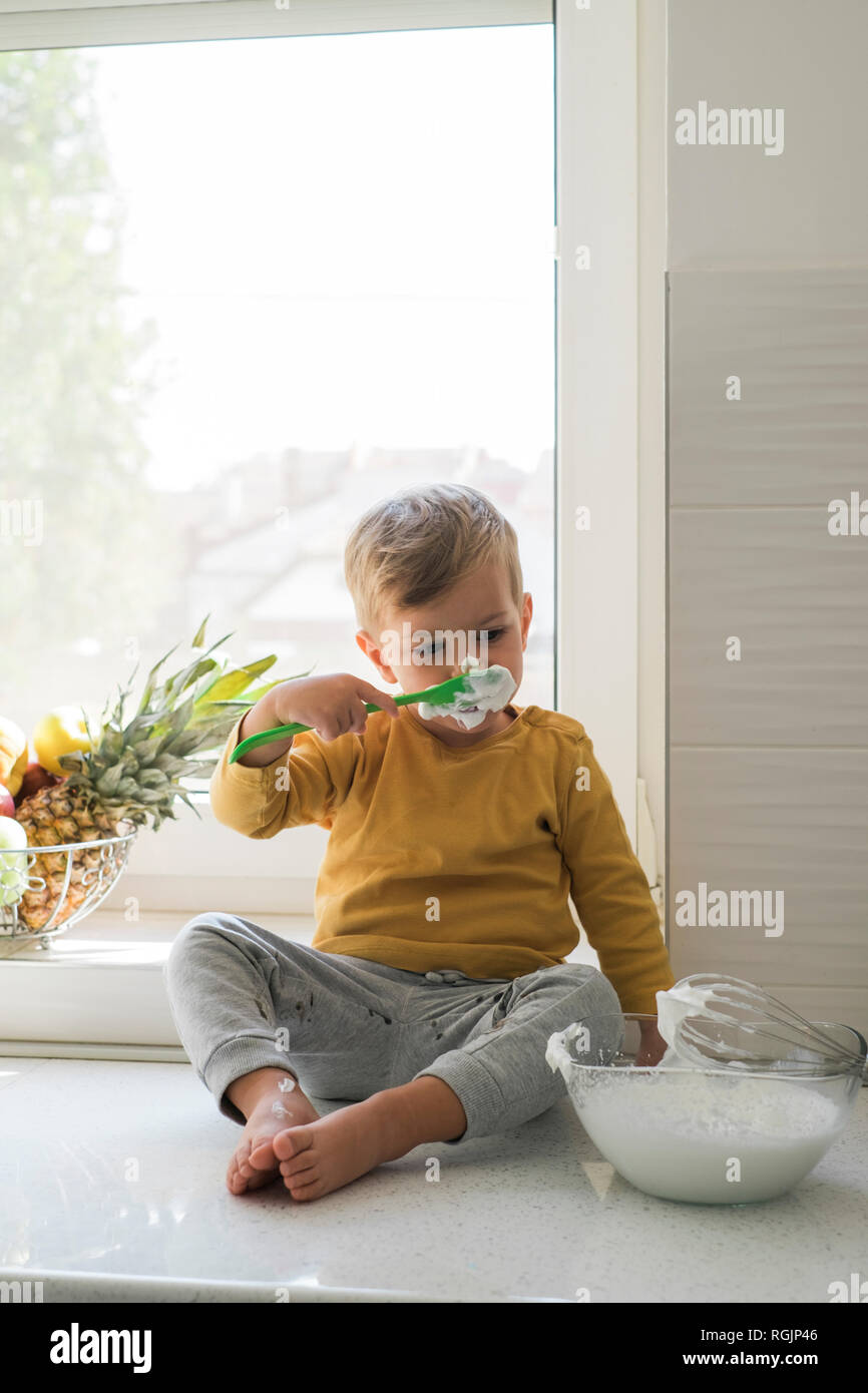 Portrait of little boy sitting barefoot n worktop in the kitchen nibbling whipped cream Stock Photo