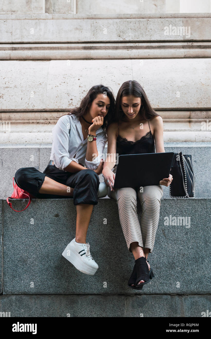 Two friends sitting side by side on a wall using laptop Stock Photo